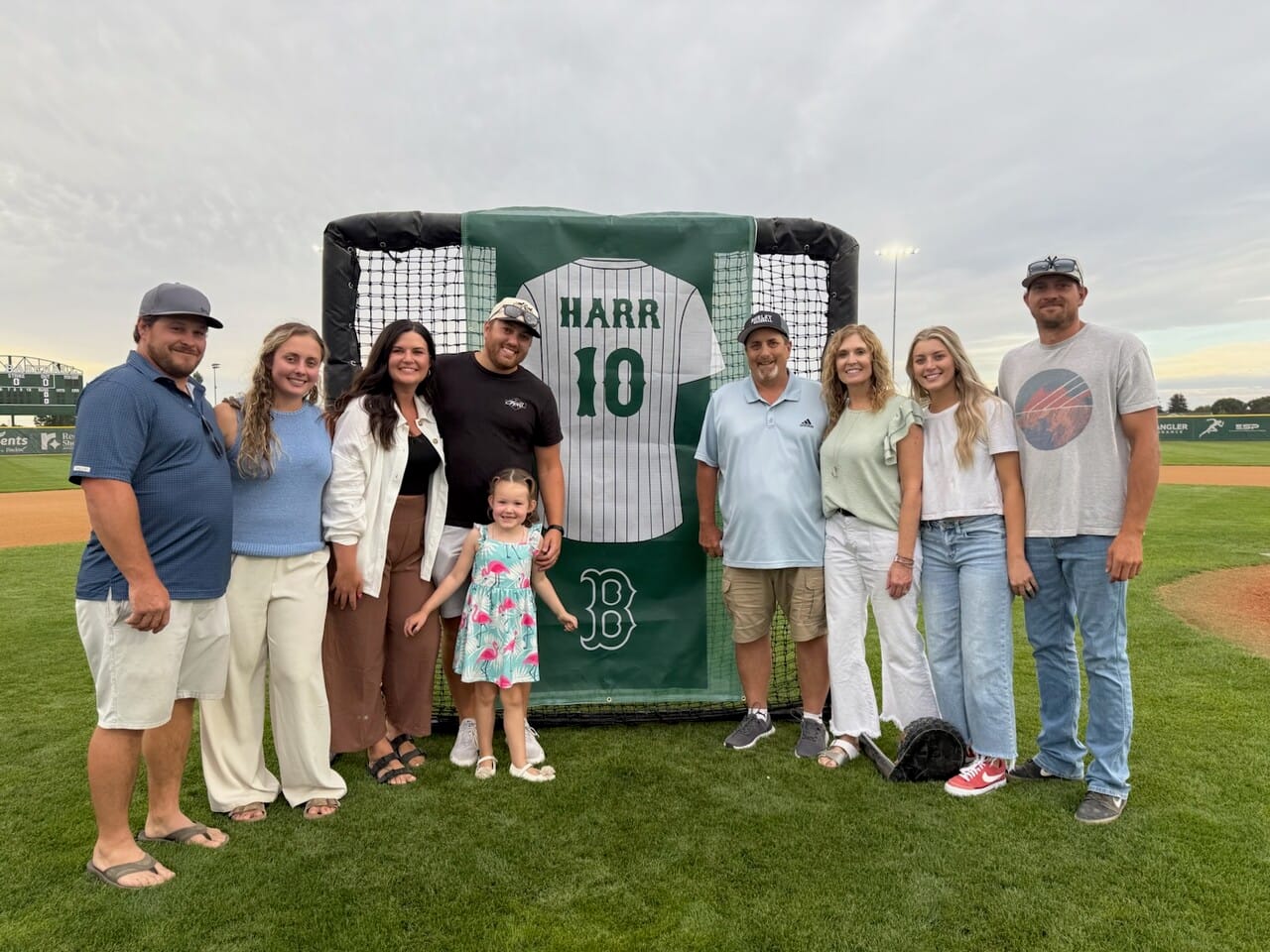 A group of adults and a young child on a baseball field, standing around a green banner of a pinstriped Harr number 10 jersey